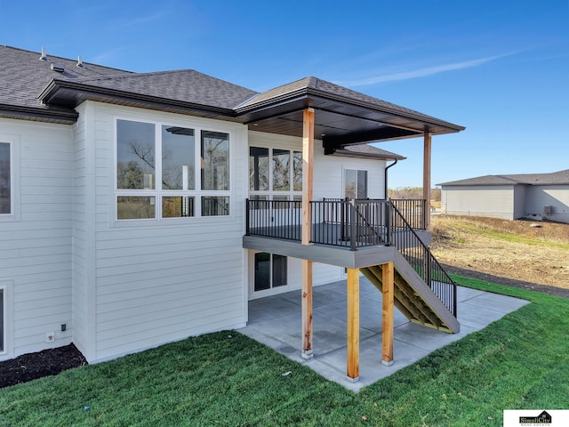 back of property featuring stairway, a wooden deck, roof with shingles, a patio area, and a lawn