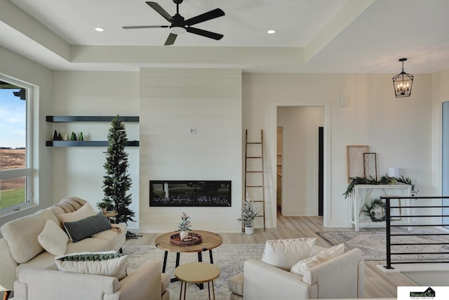 living room with a raised ceiling, wood finished floors, a large fireplace, and ceiling fan with notable chandelier