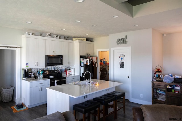 kitchen featuring a sink, washer / dryer, black appliances, dark wood-style flooring, and a kitchen bar