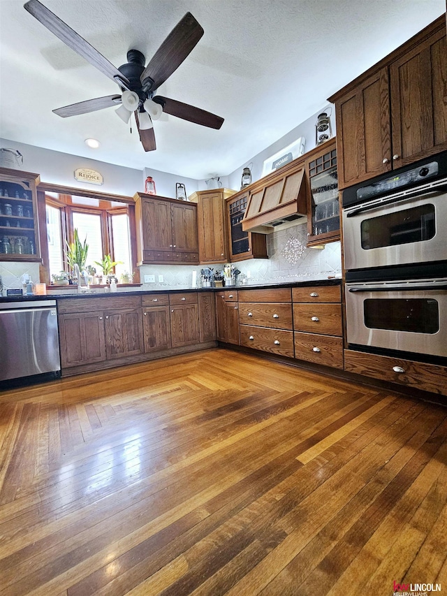 kitchen featuring glass insert cabinets, custom range hood, appliances with stainless steel finishes, dark countertops, and ceiling fan