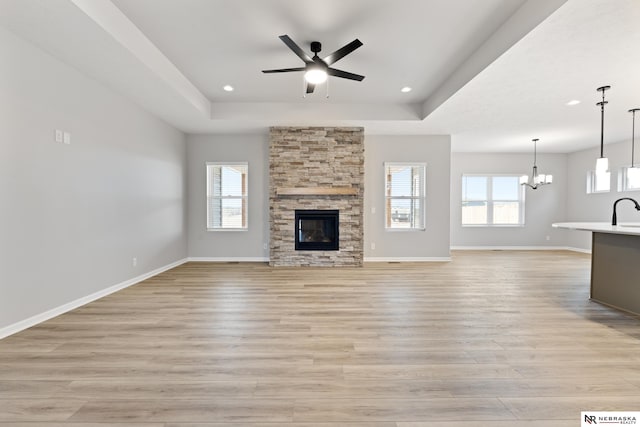 unfurnished living room featuring light wood-style floors, ceiling fan with notable chandelier, a stone fireplace, baseboards, and a tray ceiling