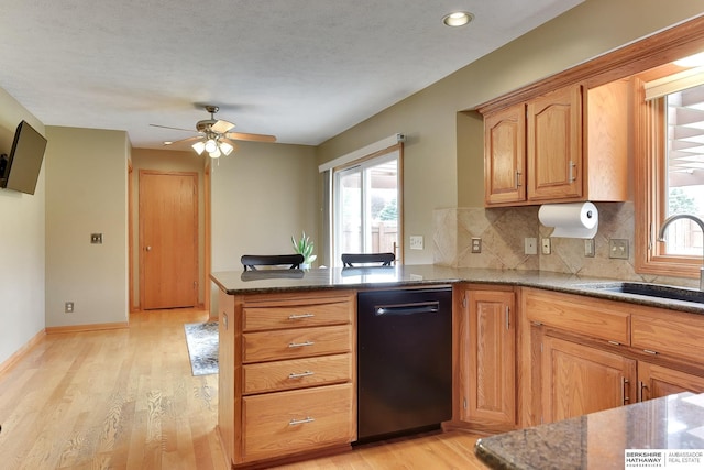 kitchen featuring a ceiling fan, dishwasher, a sink, dark stone counters, and light wood-style flooring