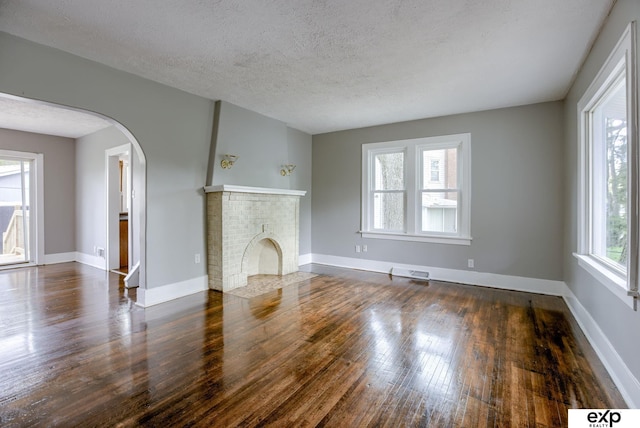 unfurnished living room featuring wood finished floors, arched walkways, a textured ceiling, baseboards, and a brick fireplace