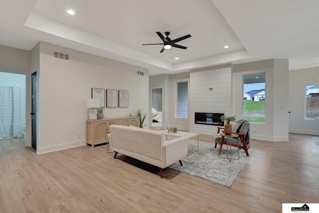 living room with visible vents, baseboards, ceiling fan, a tray ceiling, and light wood-style flooring