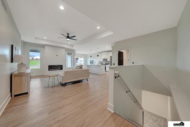 living area with ceiling fan, baseboards, light wood-style floors, a tray ceiling, and recessed lighting