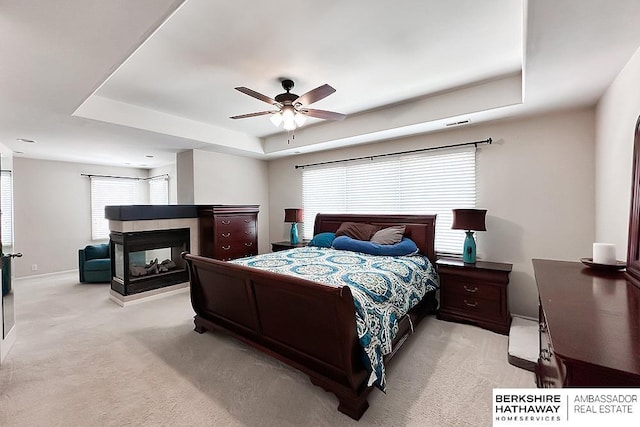 bedroom with a multi sided fireplace, a ceiling fan, visible vents, a raised ceiling, and light carpet