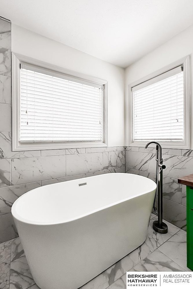 bathroom with tile walls, plenty of natural light, a soaking tub, and marble finish floor