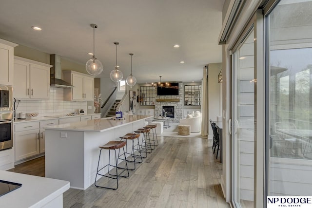 kitchen with a stone fireplace, open floor plan, light wood-style floors, wall chimney exhaust hood, and white cabinetry