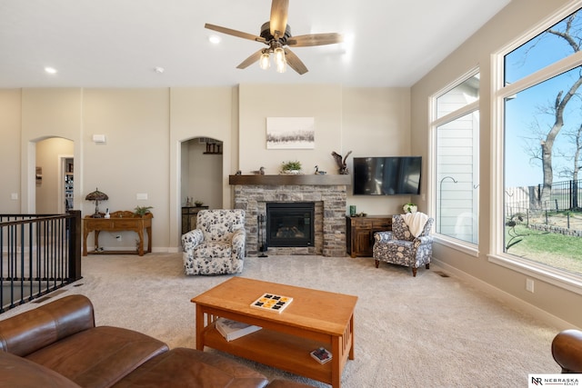 living area featuring a stone fireplace, a ceiling fan, carpet, and a healthy amount of sunlight