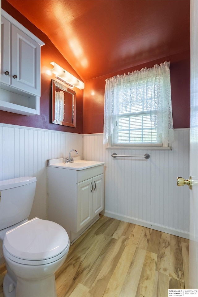 half bathroom with wainscoting, vaulted ceiling, wood finished floors, vanity, and toilet