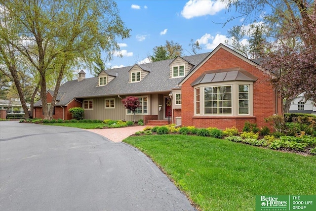 cape cod-style house with brick siding, driveway, a shingled roof, and a front lawn