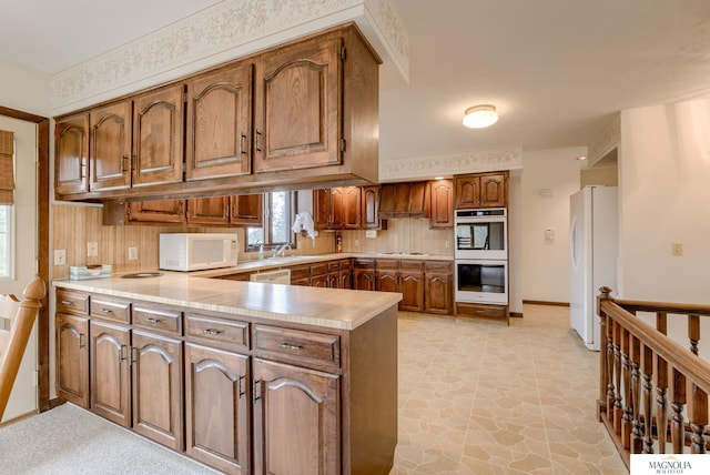 kitchen featuring a peninsula, decorative backsplash, brown cabinetry, and white appliances