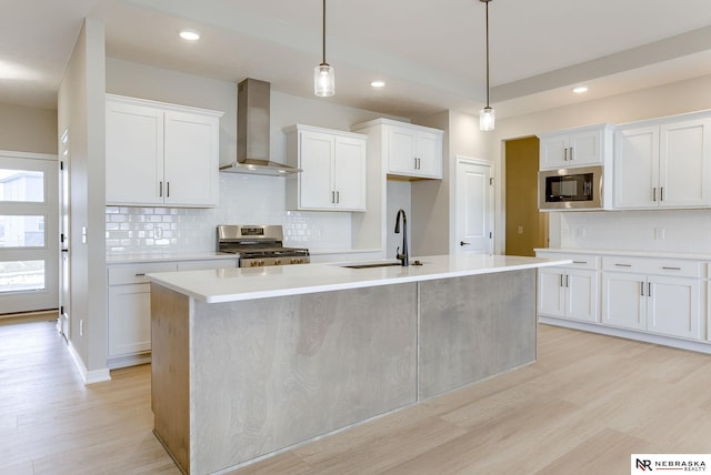 kitchen with wall chimney range hood, a kitchen island with sink, a sink, light wood-style floors, and stainless steel appliances