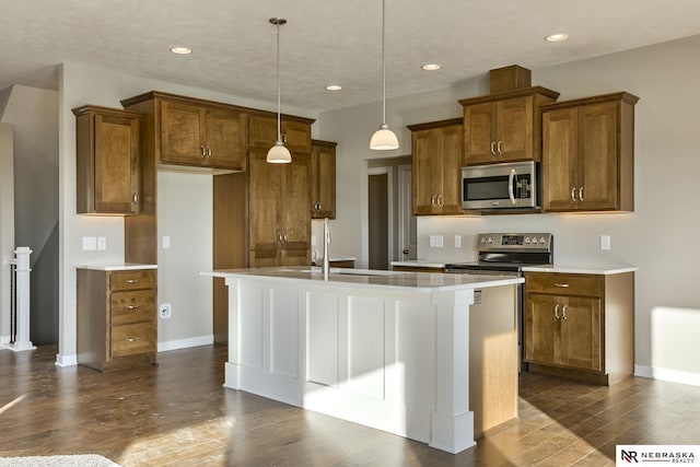 kitchen with a kitchen island with sink, dark wood finished floors, brown cabinets, baseboards, and stainless steel appliances