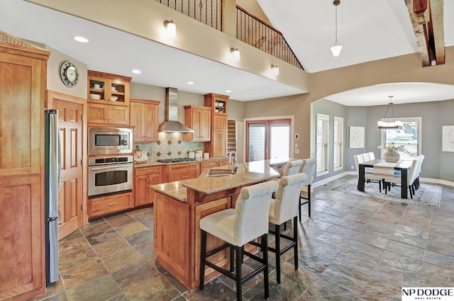 kitchen with a sink, arched walkways, wall chimney exhaust hood, stone tile floors, and stainless steel appliances