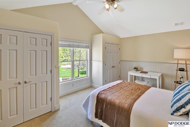 carpeted bedroom featuring ceiling fan, lofted ceiling with beams, visible vents, and a wainscoted wall