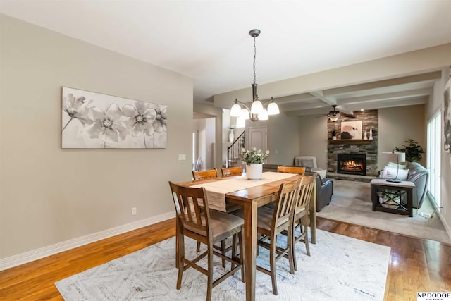 dining room featuring a fireplace, ceiling fan with notable chandelier, baseboards, wood finished floors, and beamed ceiling