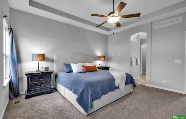 carpeted bedroom featuring ceiling fan, visible vents, arched walkways, baseboards, and a tray ceiling