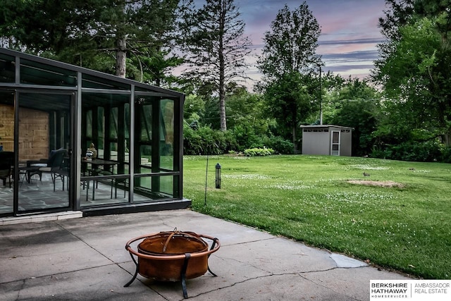 yard at dusk featuring an outdoor fire pit, a sunroom, a storage unit, a patio, and a lawn