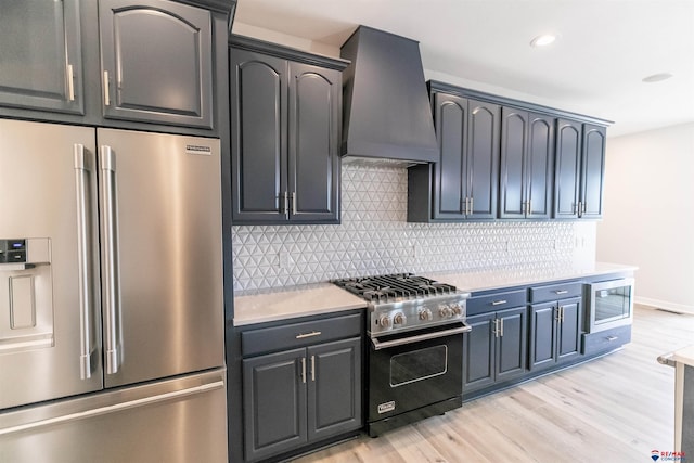 kitchen featuring high quality appliances, light countertops, premium range hood, decorative backsplash, and light wood-type flooring
