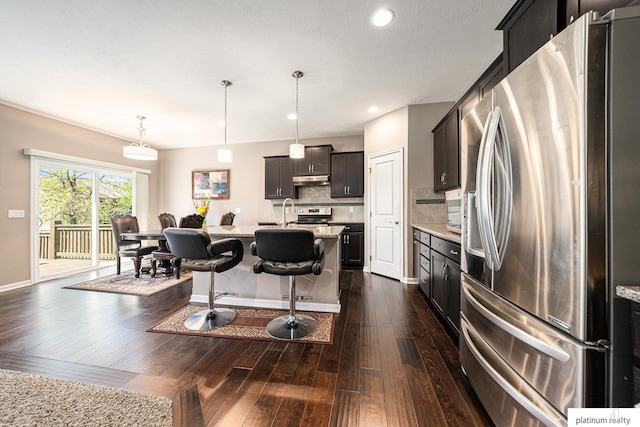 kitchen with decorative backsplash, a kitchen island with sink, a kitchen breakfast bar, dark wood-style flooring, and appliances with stainless steel finishes