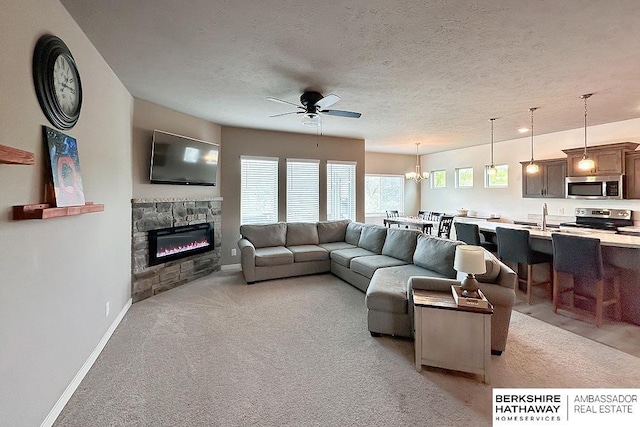 living room featuring light colored carpet, a textured ceiling, a stone fireplace, ceiling fan with notable chandelier, and baseboards