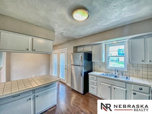 kitchen with freestanding refrigerator, tile counters, dark wood-type flooring, tasteful backsplash, and a sink