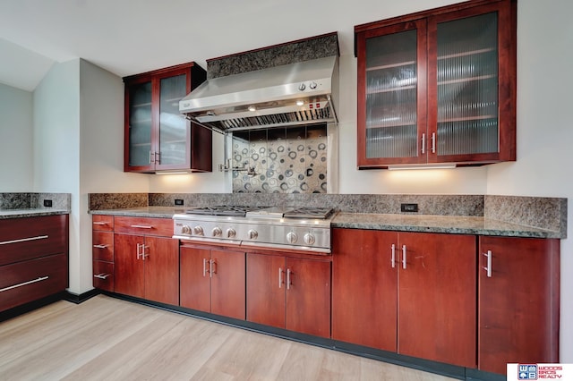 kitchen with light wood-style floors, stone countertops, wall chimney exhaust hood, stainless steel gas stovetop, and backsplash