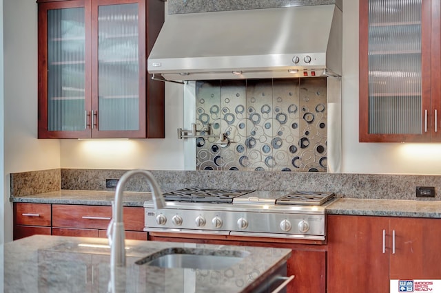 kitchen with stainless steel gas cooktop, glass insert cabinets, wall chimney range hood, brown cabinets, and light stone counters