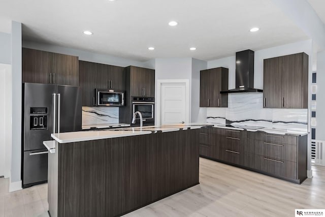 kitchen featuring wall chimney range hood, a kitchen island with sink, backsplash, modern cabinets, and black appliances