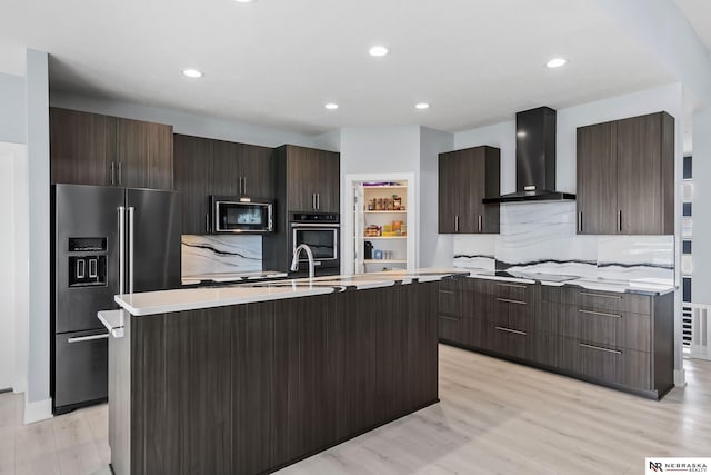 kitchen featuring modern cabinets, a center island with sink, wall chimney range hood, light countertops, and black appliances