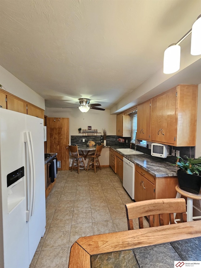 kitchen with brown cabinetry, a ceiling fan, dark countertops, a sink, and white appliances