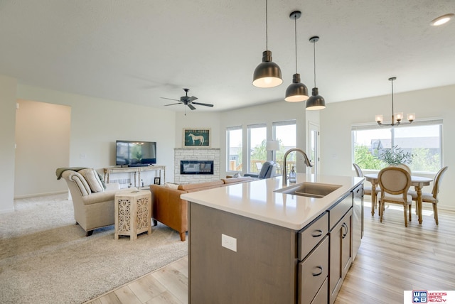 kitchen featuring a stone fireplace, a sink, ceiling fan with notable chandelier, light countertops, and an island with sink