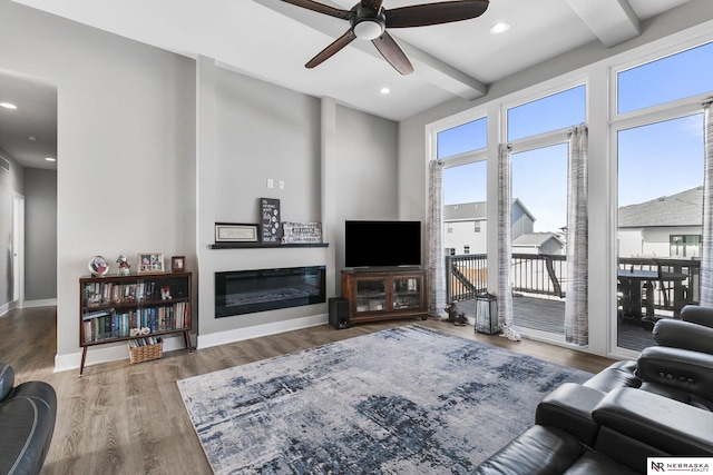 living room featuring ceiling fan, beam ceiling, a glass covered fireplace, wood finished floors, and baseboards