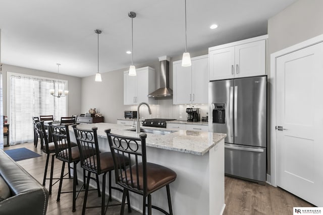 kitchen featuring stainless steel fridge, wall chimney range hood, a sink, dark wood finished floors, and a kitchen island with sink