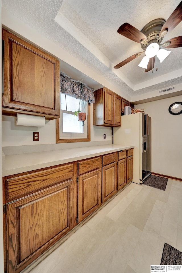 kitchen featuring brown cabinetry, a tray ceiling, light countertops, white fridge with ice dispenser, and a textured ceiling