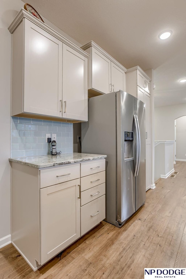 kitchen featuring light wood-style flooring, stainless steel refrigerator with ice dispenser, arched walkways, decorative backsplash, and light stone countertops