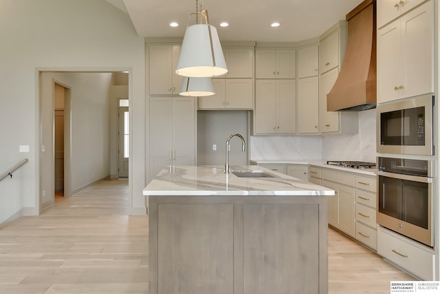 kitchen with appliances with stainless steel finishes, a sink, premium range hood, light wood-type flooring, and light stone counters