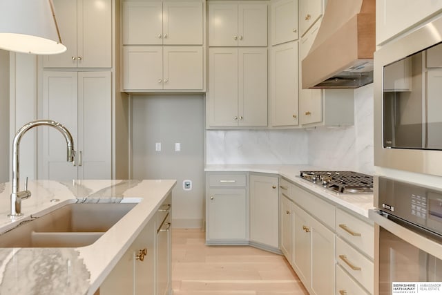 kitchen with a sink, wall chimney range hood, light wood-type flooring, backsplash, and stainless steel appliances