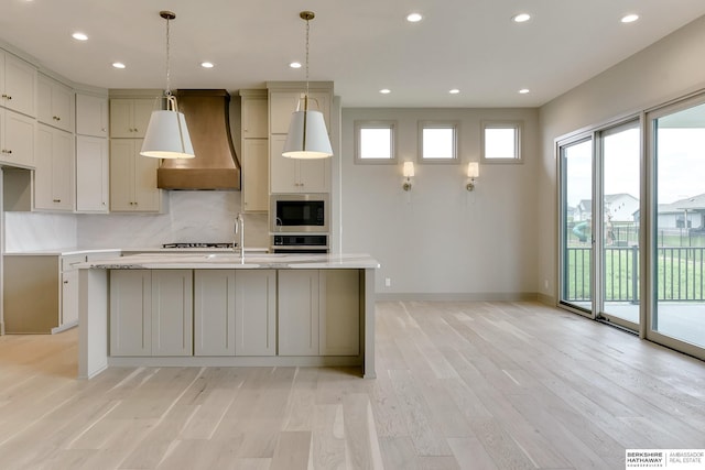 kitchen featuring wall chimney exhaust hood, built in microwave, stainless steel oven, light wood-style flooring, and a kitchen island with sink