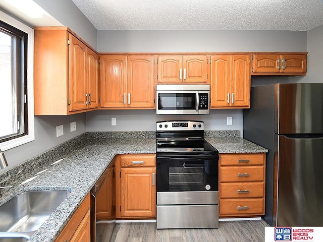 kitchen featuring appliances with stainless steel finishes, a sink, light wood finished floors, a textured ceiling, and brown cabinetry