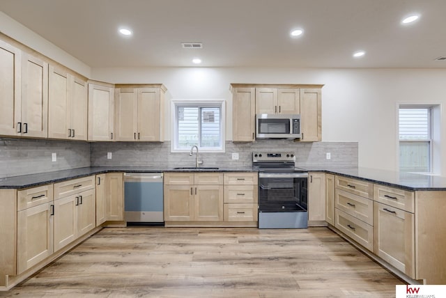 kitchen featuring stainless steel appliances, a peninsula, a sink, light brown cabinets, and dark stone counters