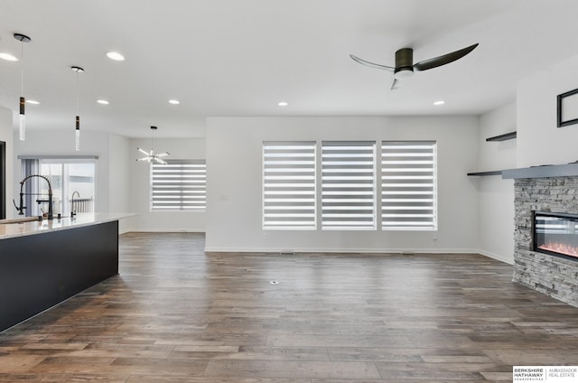 unfurnished living room featuring a stone fireplace, ceiling fan, dark wood-style floors, a sink, and recessed lighting