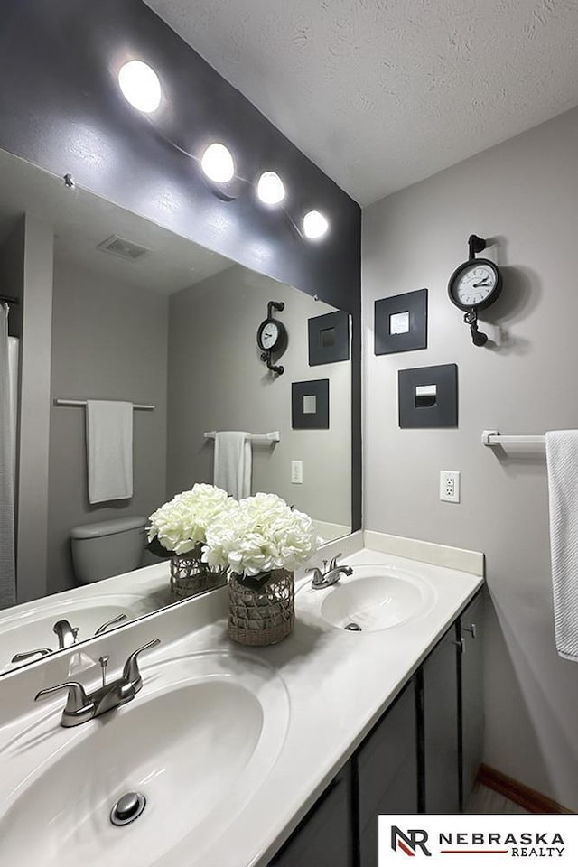full bath with toilet, double vanity, a textured ceiling, and baseboards