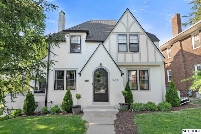 tudor house with a front yard, roof with shingles, stucco siding, and a chimney