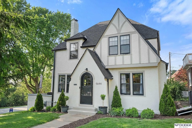 english style home with a shingled roof, stucco siding, a front yard, and a chimney