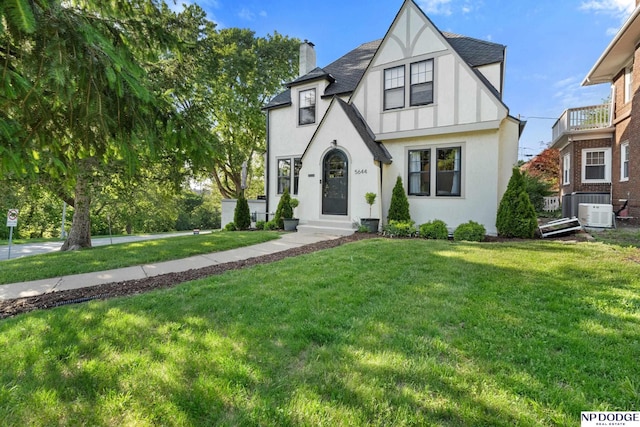 tudor home featuring a chimney, a front lawn, stucco siding, cooling unit, and roof with shingles