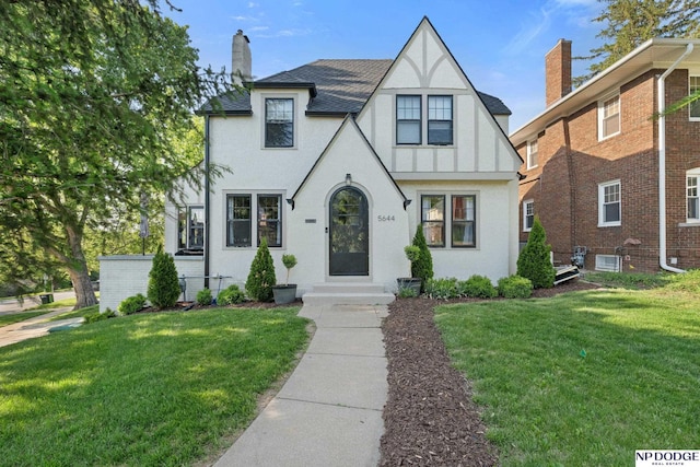 tudor home with stucco siding, a front yard, a chimney, and roof with shingles