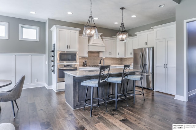 kitchen featuring stainless steel appliances, a center island with sink, a chandelier, white cabinets, and wainscoting