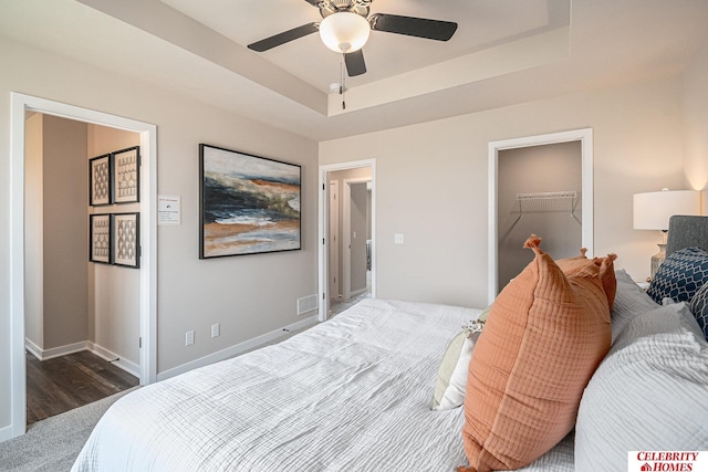 bedroom with a spacious closet, baseboards, wood finished floors, ceiling fan, and a tray ceiling